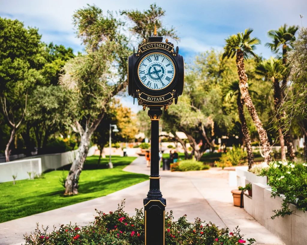 Black-and-gold vintage street clock reading "Scottsdale Rotary" surrounded by flowers, green lawn, and palm trees in a Scottsdale park, Arizona