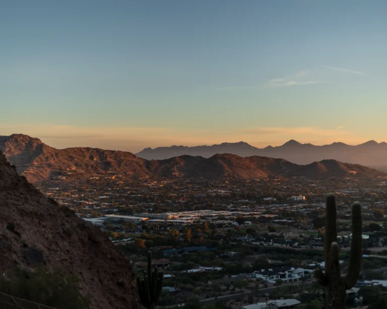 Golden-hour view from a desert mountainside — a saguaro cactus in the foreground overlooking a sprawling valley city with mountain ranges along the horizon, Arizona