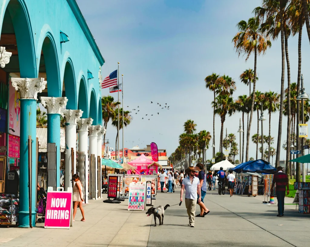 Sunny Venice Beach Boardwalk lined with palm trees, vendor stalls, and the turquoise arched colonnade building, with people and a dog walking, Los Angeles, California