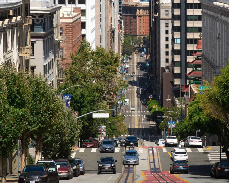 View down a steep, tree-lined San Francisco street with cable car tracks in the pavement, traffic, and high-rises — a pagoda-roofed building near Chinatown on the right, California