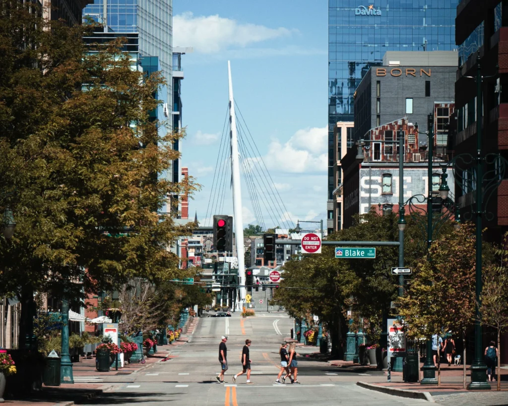 Tree-lined downtown Denver street in the LoDo district, people crossing at Blake Street with the white cable-stayed Millennium Bridge and high-rise buildings in the background