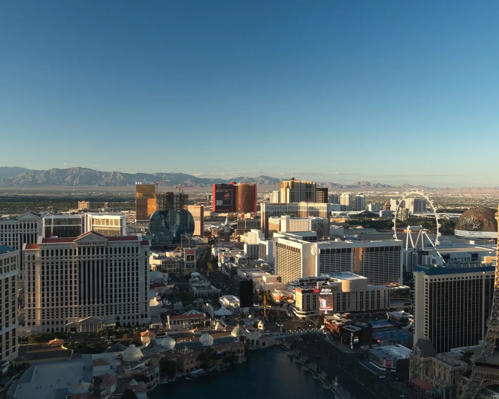 Aerial view of the Las Vegas Strip lined with resort casinos, the High Roller observation wheel on the right, and desert mountains along the horizon under a clear blue sky